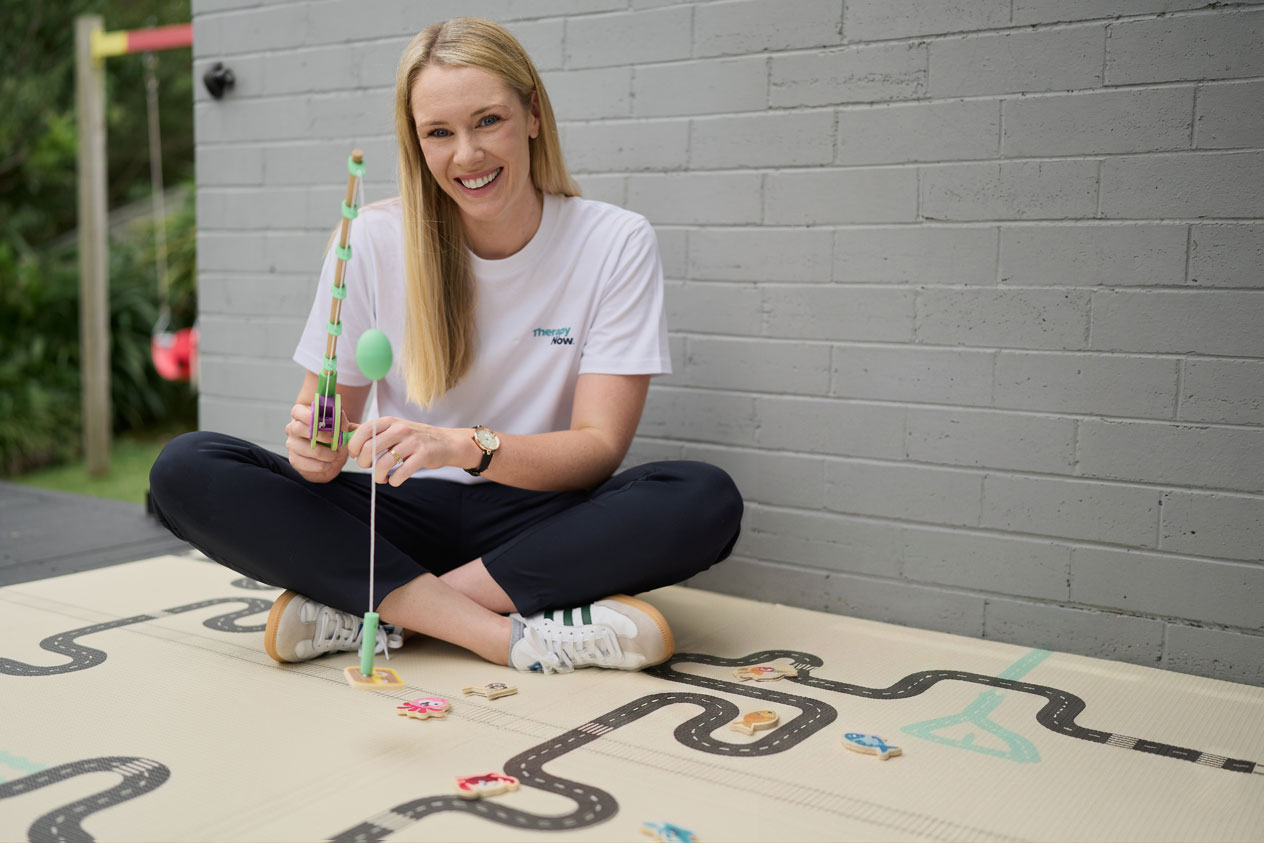 A blonde female Allied Health Assistant is sitting with her legs crossed demonstrating a magnetic fishing game for children.